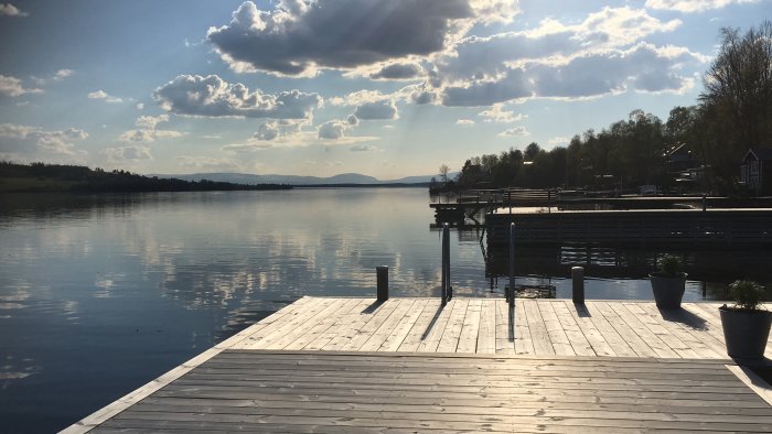 Träbrygga som leder ut till stilla vatten vid kvällstid, med Bydalsfjällen i horisonten under en molnig himmel.