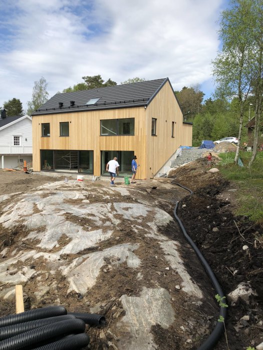 Nyanlagt dräneringsrör längs en grävd träningsränna vid ett nybyggt trähus, med två personer och byggmaterial i bakgrunden.