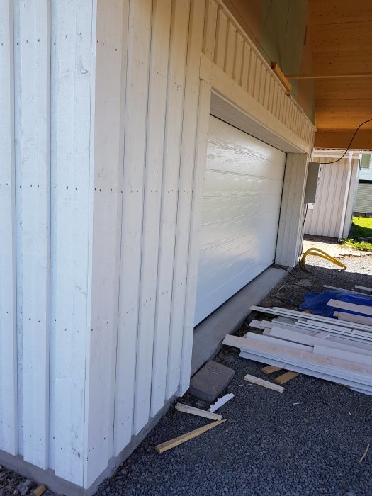 Nyinstallerad vit garageport bredvid en trävägg, omgiven av byggmaterial och brädor på marken.