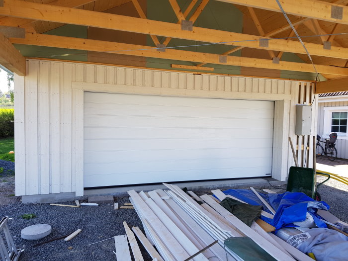 Nybyggd carport med öppet takstom och vit garageport, byggmaterial utspritt framför.