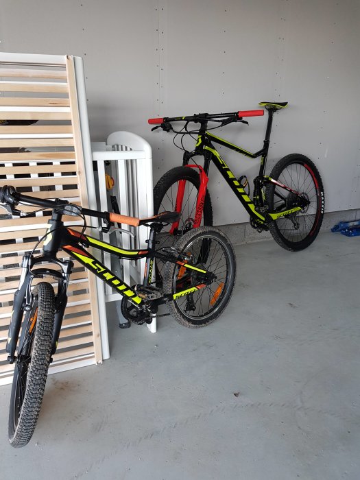 Två mountainbikes lutade mot en vägg i ett garage med grått golv och en träpall.