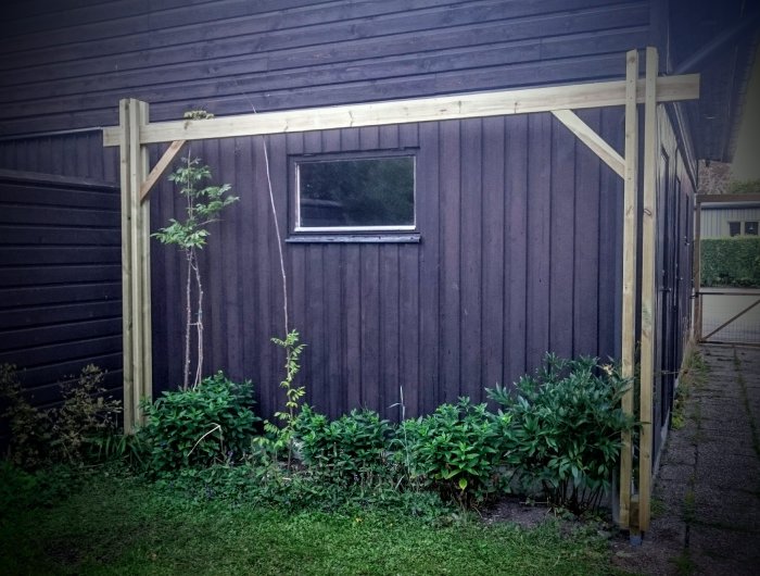 Pergola med knippepelare av reglar och distanser framför en mörk trävägg.