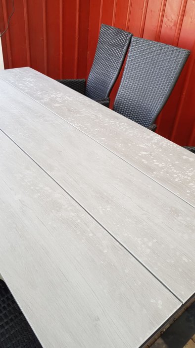 Grått konstträdäckat bord med vita fläckar efter rengöringsmedel, med två svarta vävda stolar i bakgrunden.
