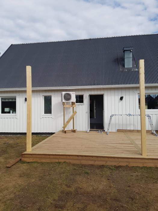 Nyinstallerat träaltangolv framför ett vitt hus med limträpelare ståendes på altangolvet och en väggmonterad AC-enhet.