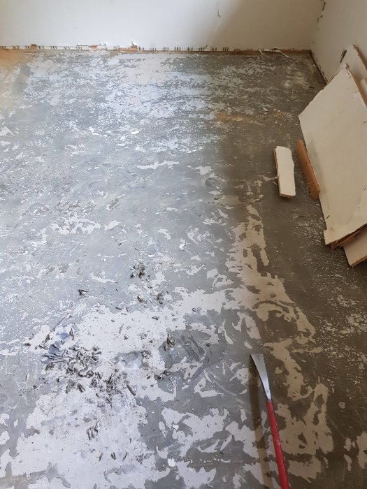 Grått betonggolv med limrester och spill efter borttagning av gammal plastmatta, inkluderar skrapa och träbitar.