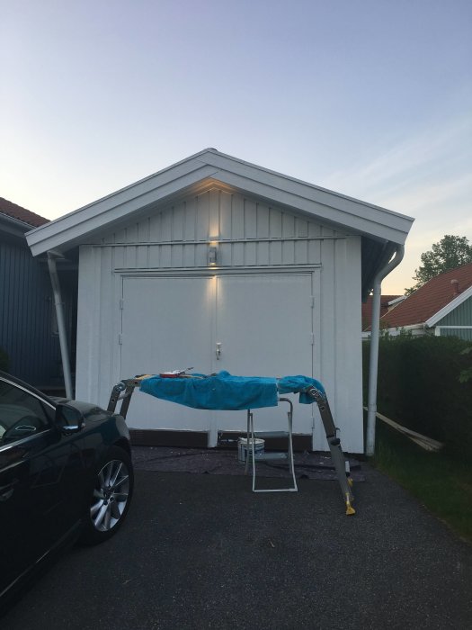 Första strykningen av vit färg på en husfasad med pensel, skyddsduk, stegar och en bil parkerad i förgrunden.