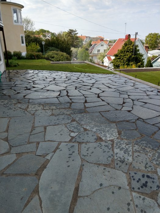 Stenlagd uppfart med oregelbundna stenplattor framför hus, gräs och hustak i bakgrunden.