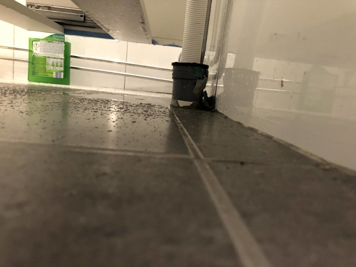 Hörn i rum med klinkergolv och vita kaklade väggar med synlig silikonfogning och en hink.