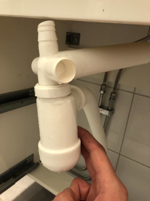 En hand som pekar på en vit avloppsfälla under en diskbänk.