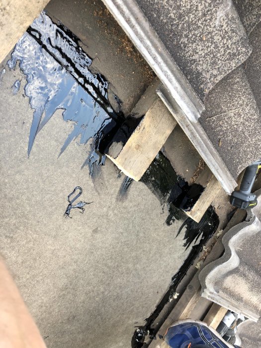 Takreparation med asfaltskitt kring ett stort hål täckt av en träskiva och betongpannor i bakgrunden.