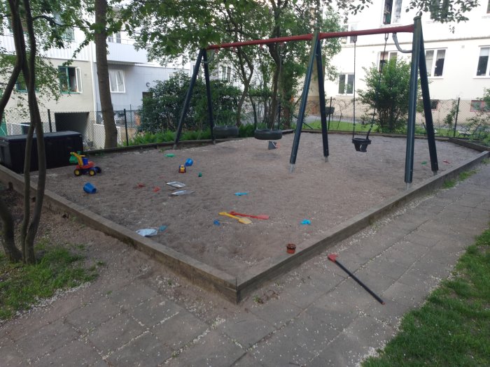 Lekplats med gungställning på sandgrund omgärdad av träreglar, leksaker utspridda på marken.