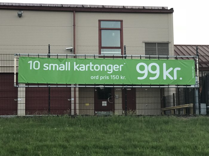 Reklambanner med texten "10 small kartonger 99 kr, ord pris 150 kr" framför en byggnad bakom stängsel.