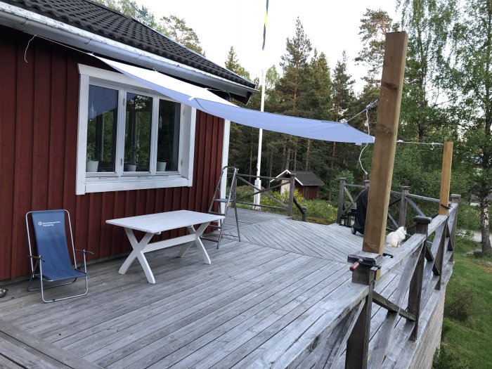 Trädäck med vit picknickbord och blå vilstol vid röd stuga, trästolpar med vit segelduk över däcket.