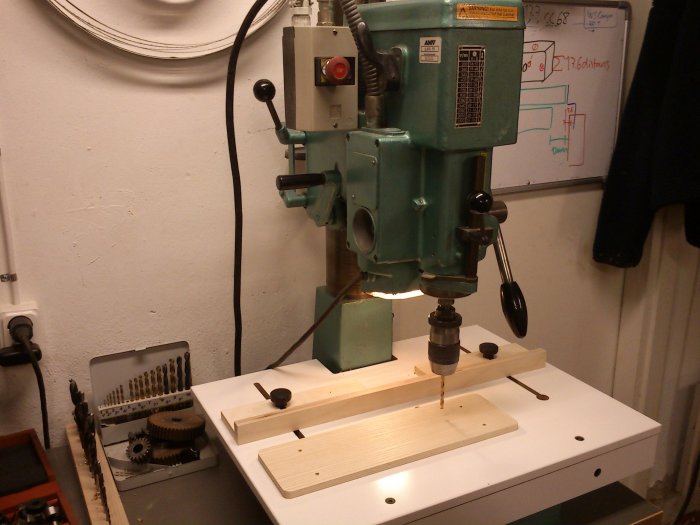 En pelarborrmaskin i arbete med träbit och anhåll med genomgående spår och L-profiler på bordet.
