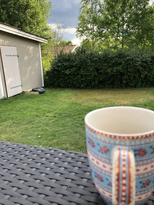 En kopp kaffe på ett trädgårdsbord med en robotgräsklippare och ett förråd i bakgrunden.