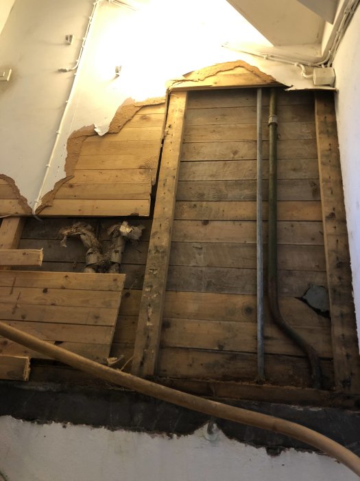 Innervägg med vattenskada och mögelpåväxt, delvis borttagen masonit och träplankor, rör till vänster.