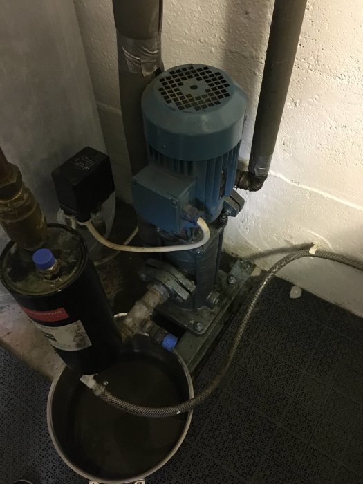 Ejektorpump och hydrofor i en källare med synliga röranslutningar och kablage, placerade på ett golv med dräneringshål.