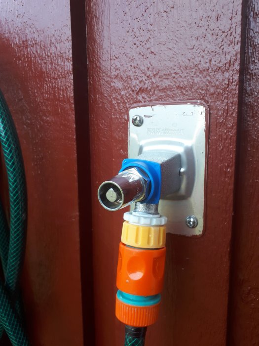 Vattenkran med kopplad slang och fyrkantig krananslutning mot en röd trädörr.