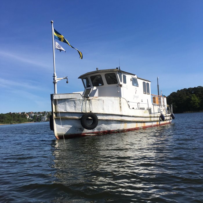 Båt för ankar i vatten med svensk flagg, nära en huvudstad, med svagt synlig kustlinje i bakgrunden.