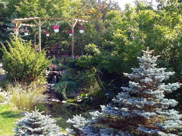 Pergola med 70x70 mm stolpar över en 8-kantig altan vid en damm, omgiven av grönska och blommor.