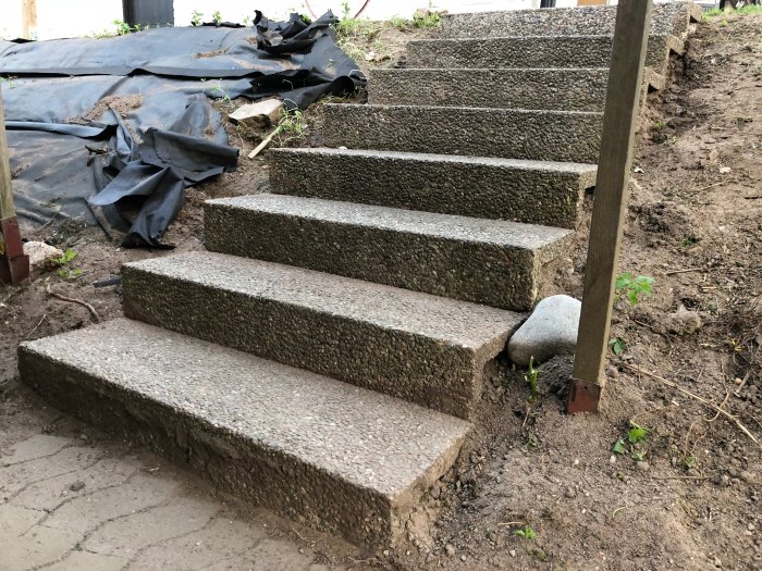 Nybyggd trappa utomhus med fyra betongsteg, kantad av en jordig trädgård och en svart presenning i bakgrunden.