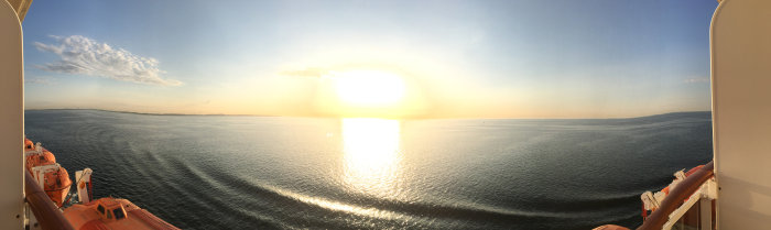 Panoramabild tagen från ett fartyg med solnedgång över havet och livbåtar på sidan.
