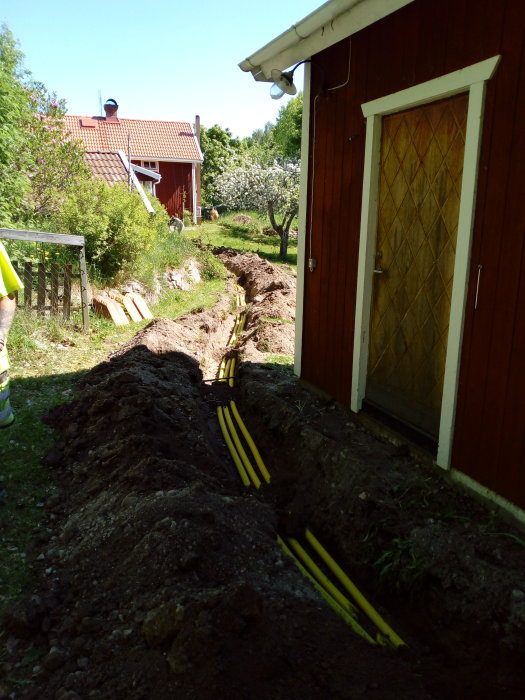 Grävd schakt i trädgård med nya gula rör för el och fiber bredvid en röd byggnad och grönska.