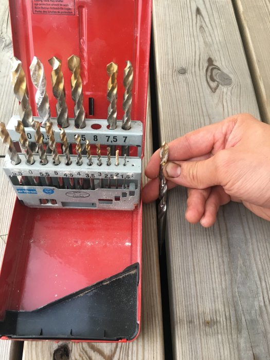 Hand som håller i en 8 mm borr bredvid en röd borrbox ovanför en trall med märkbara krympningsgap.