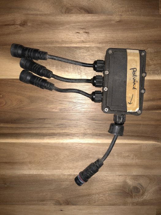 Förgreningsbox med tre utgångar på trägolv, märkt "Provning" och en ingång, relaterad till altanbelysning.