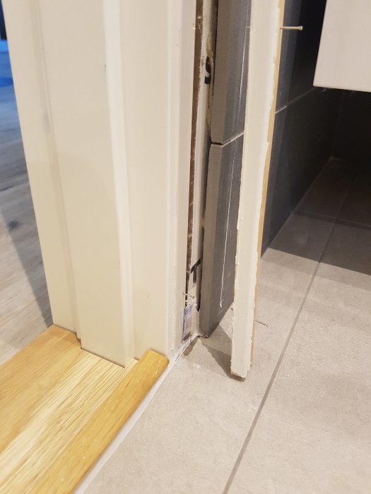 Delvis demonterad dörrkarm och tröskel som visar skadad vägg och golv-underlag.