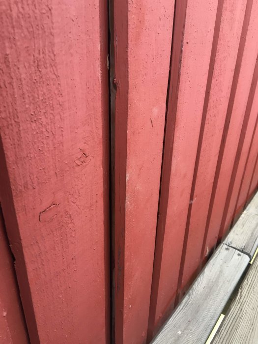 Rödmålad träfasad med sprickor i färgen och en snedspikad panelbräda, indikationer på behov av underhåll.