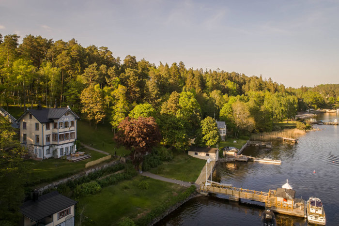 Vy över en stor, klassisk villa vid vattnet omgiven av gröna träd och en brygga med båtar.