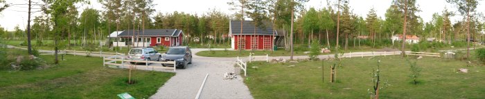 Panoramavy över ett nybyggt staket i en trädgård, vitmålade stolpar och slanor, med bakgrund av röda hus och gröna träd.