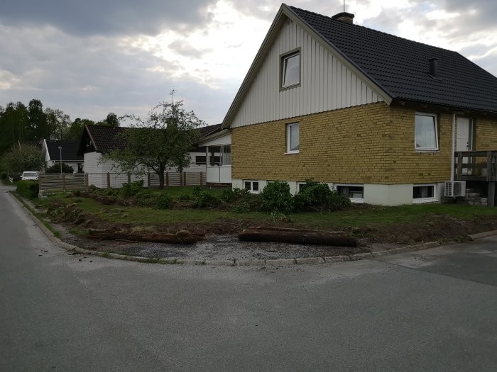 Hörntomt med grävd mark längs trottoaren där en stenmur ska anläggas, huset i bakgrunden.