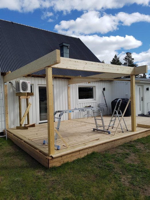 Halvfärdigt trädäck med löst liggande trall och ett påbörjat stolptakstomme framför ett hus med sadeltak.