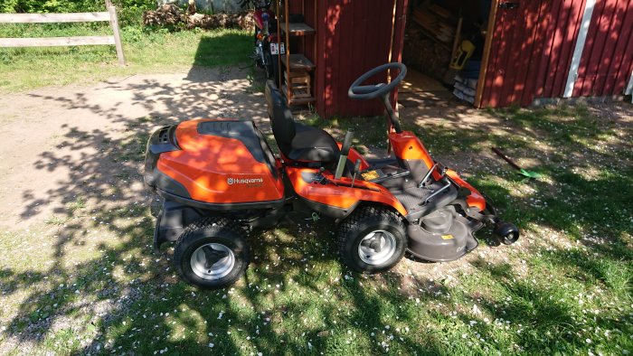 Orange Husqvarna åkgräsklippare parkerad utomhus framför en röd bod.