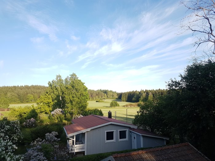 Utsikt över nyrenoverat grått hus med rött tak och grönskande trädgård mot landskap med fält och skog.