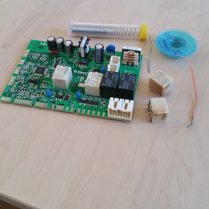 Kretskort från diskmaskin med två utbytta reläer, lödtråd, desoldering wick på ett träbord.