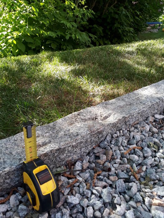 Måttband visar höjden på granitsten jämfört med grus och gräs i trädgårdsmiljö.