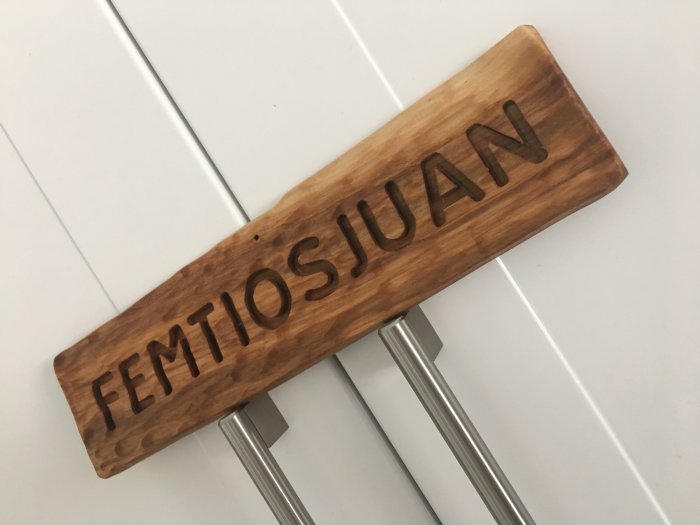 Handbearbetad träskylt med texten "FEMTIOSJUAN" ingraverat i trafikverkets typsnitt, okänt trädslag.