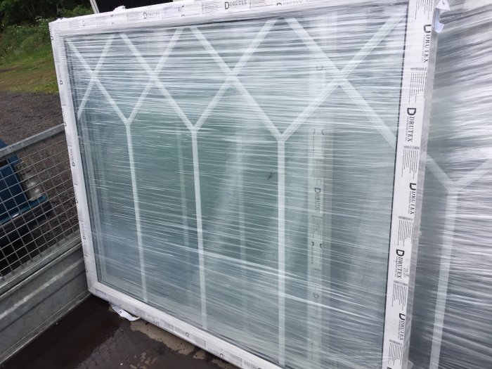 Nya Drutex PVC-fönster med invändig spröjs, plasten kvar, staplade utomhus.
