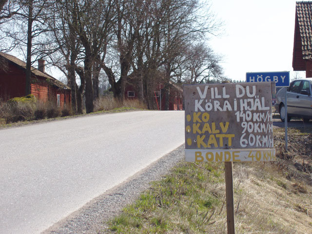 Skylt med texten "VILL DU KÖRA IHJÄL: KO 110KM/H, KALV 90KM/H, KATT 60KM/H, BONDE 40KM/H" vid lantväg.