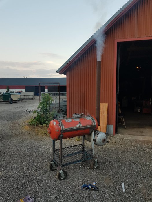 Rök kommer ut från en hemmagjord röd grill placerad utomhus nära en lada.