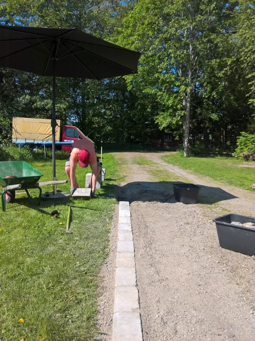 Tonåring arbetar med att lägga marksten på en gångväg i en trädgård med grönytor och träd i bakgrunden.