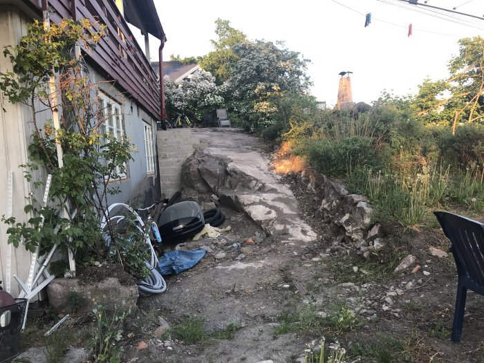 Rörig trädgård med stenigt bergsparti vid hus, potentiell plats för trappbygge mellan två nivåer.