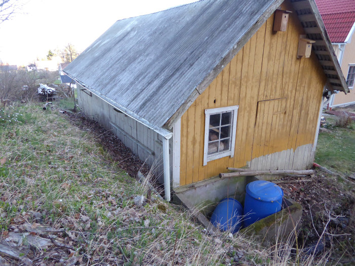 Gammalt gulmålat garage med slitet tak och byggnadsavfall runtom, beläget i naturlig sluttning.