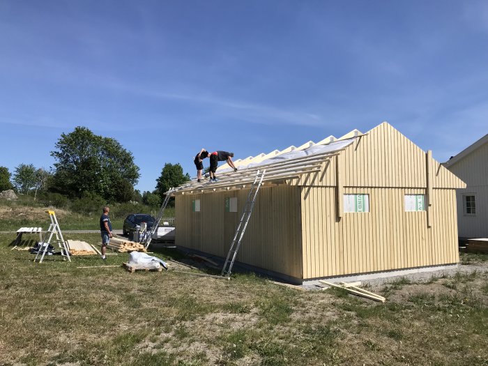 Arbetslag monterar takstolar på en träbyggnad under konstruktion en solig dag.