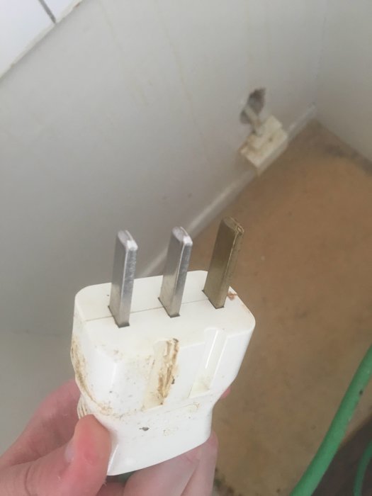 Hand håller en smutsig vit spisplugg med tre metallstift, ett märkt med jordtecken, mot en vit vägg.