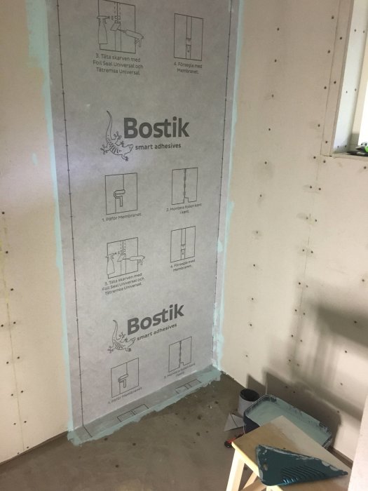 Vägg i badrum som delvis täckts med Bostik tätskikt och instruktioner, bredvid en hink och spackelspade.