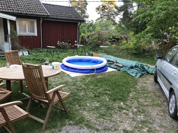 Trädgård framför rött hus med lågt tak, möbler, uppblåsbar pool och parkerad bil.